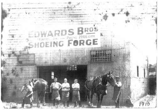 Three men in work aprons pose in front of the open doorway of a large tin shed, Edwards Bros Shoeing Forge. A fourth man on the right is holding a horse buy the bridle while a fifth man on the left is holding the back left leg of a second horse a hammer in his right hand apparently replacing a horse shoe.