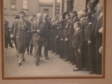 Several men in suits, some with medals line up in front of a building while a man and women followed by several other people pass by. Two men are doffing their hats as the group pass by.