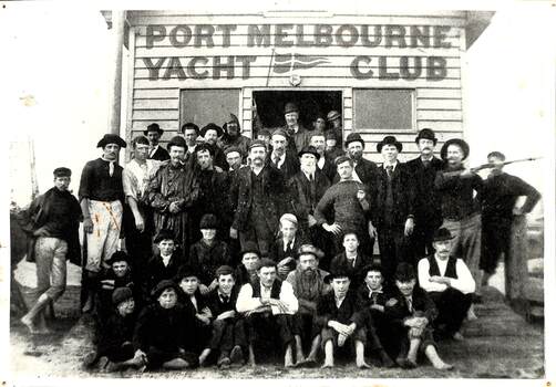 Black and white image of a group of men and boys posing outside a small wooden building identified as Port Melbourne Yacht Club.