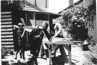 Three boys in the back yard of a house. One carries a piece of timber on his shoulder while the other two are hammering timber on a bench.
