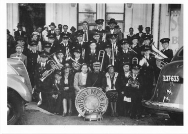 A large group of people, some in uniform with musical instruments, pose in front of a building. A drum in front of the group reads 'Port Melbourne Boys Band'.