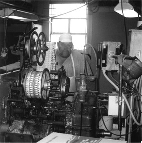 A man in a white cap stands behind a piece of machinery on a bench.
