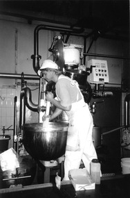 A man in white cap and overalls stirs the contents of a large cauldron.