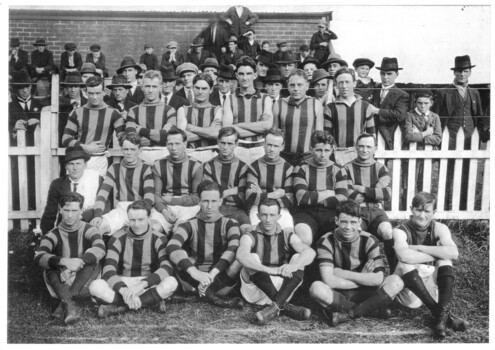 A group of men in sports uniform posing in three rows in front of a picket fence with many people in the background on the other side of the fence.