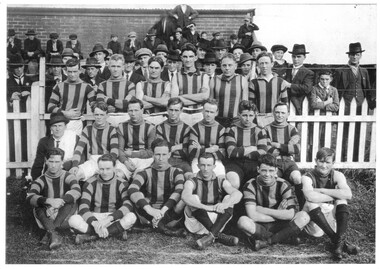 A group of men in sports uniform posing in three rows in front of a picket fence with many people in the background on the other side of the fence.