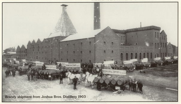 Many people pose with many horse-drawn carts loaded with barrels in from of an industrial building.