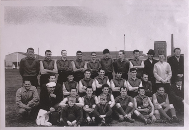 Football team including officials, training staff and three mascots (two boys and a baby) posing in three rows. Front row sitting on the ground, second row seated or crouched and the back row sitting.