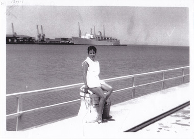 A woman poses, sitting on a bollard on a pier with a cruise ship berthed at another pier in the background.