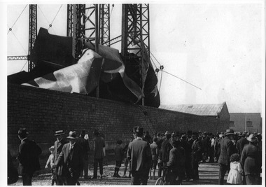 People gather in a street separated from a crumpled gasometer by a high wall.