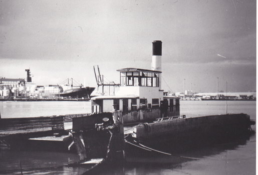 Black & white photo showing a ferry docked on a river, with other ships and building in the background.