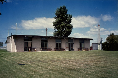 Single storey building with five sets of double doors or windows facing an open grassed area. Several park benches sit in front of the building facing the grassed area.