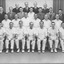 A group of twenty-six men pose in typical sporting club style in four rows. Fourteen men, mostly in the front two rows, are wearing cricket whites, the remaining men are wearing suits and ties.