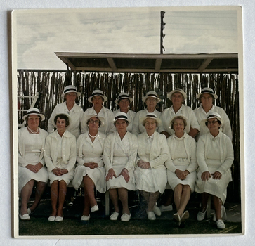 Thirteen women in white uniforms including white hats and shoes, seven seated in the front row and six standing behind.