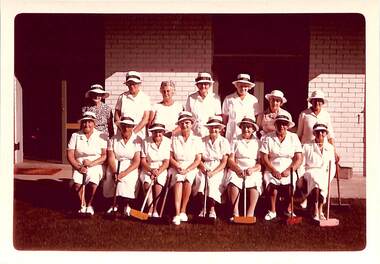Fifteen women, mostly in white uniforms and hats, pose in team format holding large wooden mallets. Seven women standing behind eight women seated. 