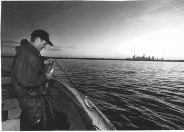 A man standing in a boat holding a fishing net containing some small fish. A city skyline is visible across a large stretch of water.