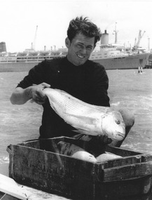 A man holding a large fish on a boat or onshore. Over a stretch of sea in the background a ship is berthed.