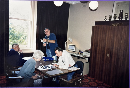 Three people sitting around a table, two are studying a group of photographs. A fourth person is standing near the table examining a glass bottle.