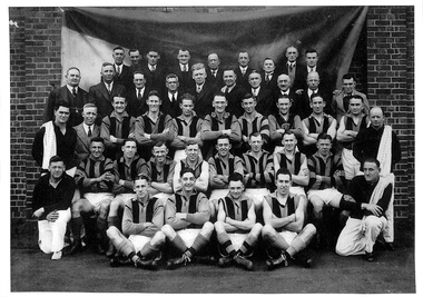 Five rows of men in typical sporting team pose. Men in two back rows wearing suits with the remaining three rows a mix of players, trainers and other staff