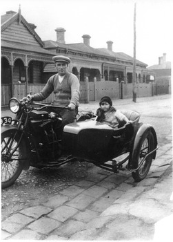 A man on a motorbike in a flat cap with a young girl wearing a coat, scarf and hat in the sidecar.