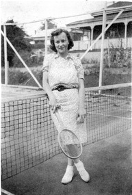 A women poses on a tennis court, next to the net, arms by her side, holding a tennis racket in her right hand and a tennis ball in her left.