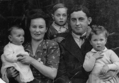 A woman and man pose for the camera. The woman is holding a toddler, a second child peers from behind and between the adults and the man has a young girl sitting on his knee.