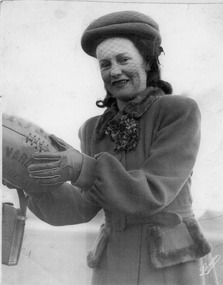 A stylishly dressed woman seen from the waist up, in a hat, with a net veil over her face, and wearing gloves smiles at the camera while holding an Australian Rules football.