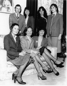 Three women pose sitting on a couch while a man and three other women pose standing behind the couch.