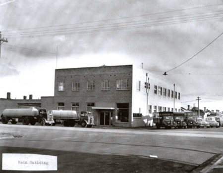 A substantial two storey brick building on a street corner. Two milk tankers are parked parallel to the front of the building and several vans and cars are reverse-parked on an angle along the side of the building.