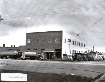 A substantial two storey brick building on a street corner. Two milk tankers are parked parallel to the front of the building and several vans and cars are reverse-parked on an angle along the side of the building.