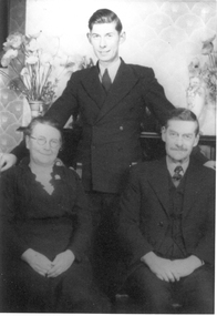 A young man in a suit and tie stands behind an older seated couple. His right hand is resting on the women's right shoulder and his left hand is resting on the man's left shoulder. The woman is wearing a black dress and round glasses. The man is wearing a suit, waistcoat and tie and has a moustache.