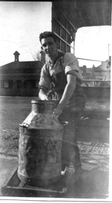 A man in shirt sleeves and overalls standing behind a milk churn.