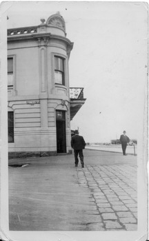 A man is walking away from the camera, approaching the corner entrance to a two storey Victorian-style hotel. Another man is standing a little distance away with his back to the hotel.