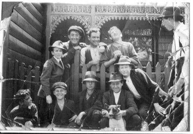 An informal portrait of a group of ten men and boys and a dog either side of a wooden picket fence in front of a house.