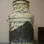 A metal milk churn that has been painted with coloured flowers on a cream background. A lot of the paint has peeled of but 'WOODRUF' and 'D A I' is still visible.