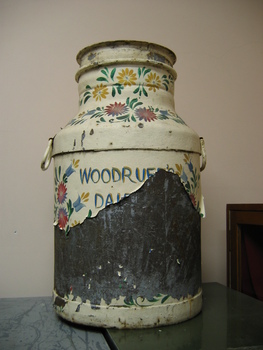 A metal milk churn that has been painted with coloured flowers on a cream background. A lot of the paint has peeled of but 'WOODRUF' and 'D A I' is still visible.