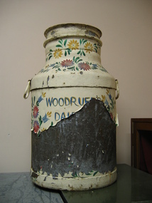 The metal milk can that has been painted with coloured flowers on a cream background. A lot of the paint has peeled of but 'WOODRUF' and 'D A I' is still visible.
