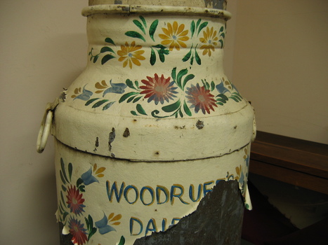 Closeup of the top half of a milk churn painted with colourful flowers on a cream background. The paint on the lower part of the churn is peeling badly but most of the name "Woodruff's Dairy" is still visible.