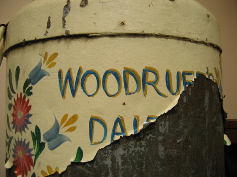 Closeup of what remains of the name "Woodruff's Dairy" on a milk churn painted with colourful flowers on a cream background. The paint has peeled badly so the last few letters of each word are no longer visible.