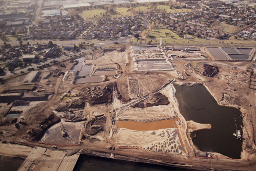 Aerial view of a large area of wasteland adjoining a housing estate. The wasteland has signs of work with several manmade depressions, both regular and irregular, filled with water.