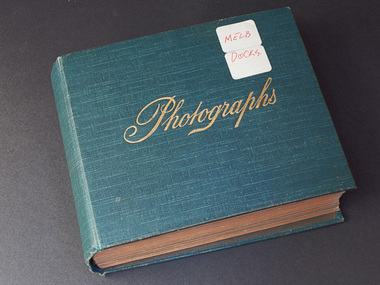 A thick photograph album with a blue cover with two small white stickers with "MELB" on one and "DOCKS" on the other handwritten in red 