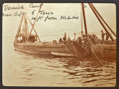 Two work boats bow to bow at the end of a small wharf. One of the boats has a block and tackle and several men spread between the two boats and the wharf are working to lift some timber from the water.