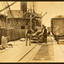 Several workmen stand on a pier or wharf next to a pile of sacks their backs to the camera. There is a railway wagon to their right full of sacks and a ship berthed to their left.