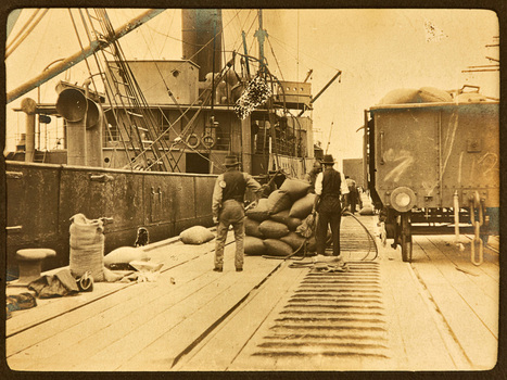 Several workmen stand on a pier or wharf next to a pile of sacks their backs to the camera. There is a railway wagon to their right full of sacks and a ship berthed to their left.