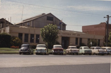 Coloured photo of buildings with early make parked on roadside. 