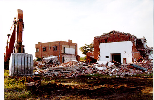 An earthmover sits idle in the left foreground beside an area of rubble and two partly demolished buildings in the background.