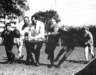 Two men in everyday clothing playing football as five boys attempt to tackle them.