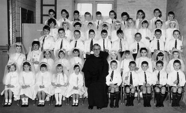 Black & white photo of children who had received their 1st communion standing with a priest. 