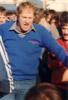 A man in a blue jumper and jeans, his mouth wide open, is hemmed in by a crowd of people behind him. 