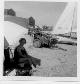 Elizabeth Cannatelli sits on the beach in the shade of a large umbrella. A car trailer and a sailing dingy, with sails raised are visible on the beach behind her.