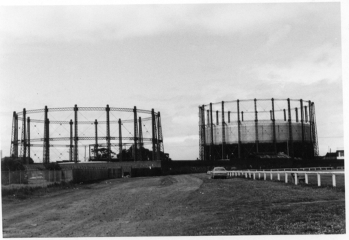 Black & white photo of two large gasometers with large open space in front of them. 